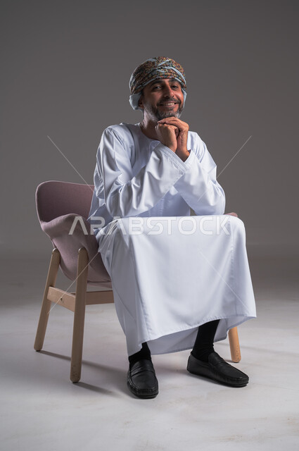 التأمل والتخطيط للمستقبل، النظر الى الكاميرا بتعابير الفرح والسرور، بورتريه لرجل عربي خليجي عماني مبتسم يرتدي الدشداشه والعمامة يجلس على كرسي مريح ويضع يديه تحت ذقنه، إيجاد حل لمشكلة ما واتخاذ القرارات، خلفية رماديه