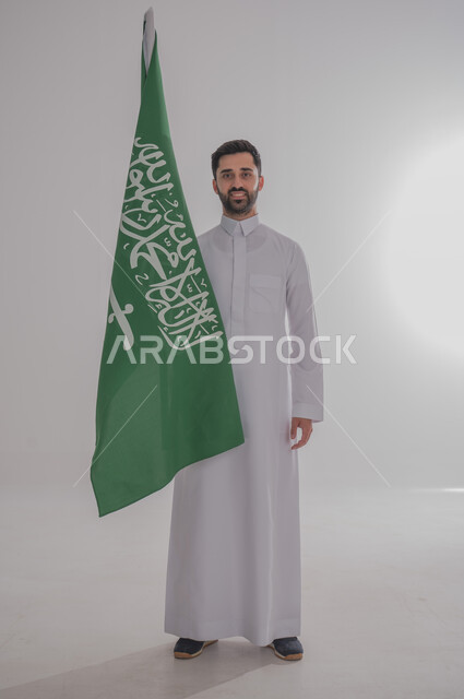 Participating in national occasions and holidays, the concept of love and belonging to the homeland, the memory of Saudi National Day, a portrait of a Saudi Gulf Arab man wearing a dishdasha holding the flag of the Kingdom of Saudi Arabia in his hand, celebrating the Kingdom's National Day, gray background