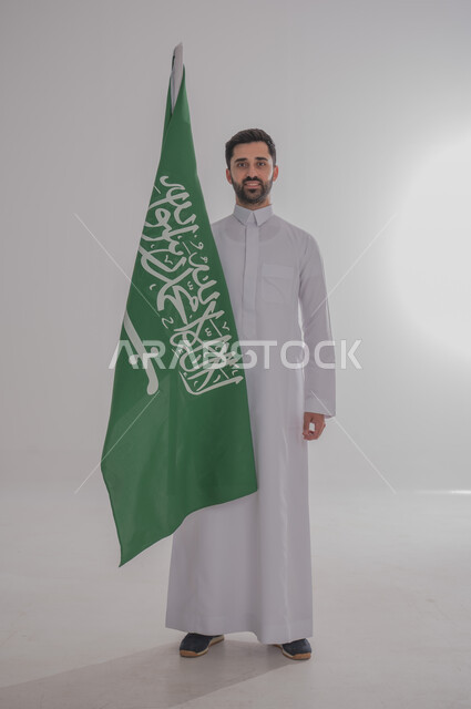 Participating in national occasions and holidays, the concept of love and belonging to the homeland, the memory of Saudi National Day, a portrait of a Saudi Gulf Arab man wearing a dishdasha holding the flag of the Kingdom of Saudi Arabia in his hand, celebrating the Kingdom's National Day, gray background