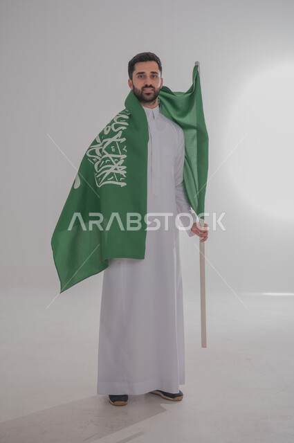 Participating in national occasions and holidays, the concept of love and belonging to the homeland, the memory of Saudi National Day, a portrait of a Saudi Gulf Arab man wearing a dishdasha and placing the flag of the Kingdom of Saudi Arabia on his shoulder, celebrating the Kingdom's National Day, gray background