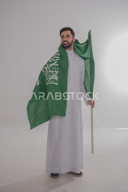 المشاركة بالمناسبات والاعياد الوطنية، مفهوم حب الوطن والانتماء له، ذكرى اليوم الوطني السعودي، بورتريه لرجل عربي خليجي سعودي يرتدي الدشداشة يضع على كتفه علم المملكة العربية السعودية، الاحتفال بالعيد الوطني للمملكة، خلفية رمادية