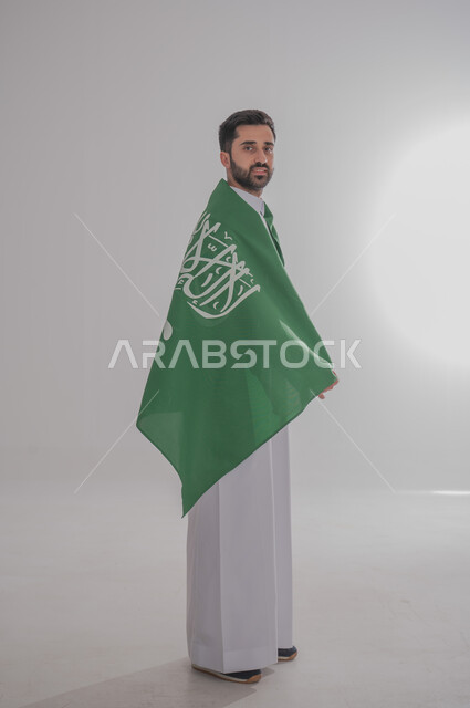 Participating in national occasions and holidays, the concept of love and belonging to the homeland, the memory of Saudi National Day, a portrait of a Saudi Gulf Arab man wearing a dishdasha and placing the flag of the Kingdom of Saudi Arabia on his shoulder, celebrating the Kingdom's National Day, gray background