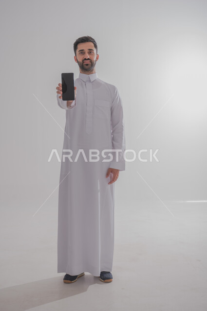 التواصل مع الأهل والأصدقاء عبر الهاتف المحمول، استخدام الأجهزة الحديثة والمتطورة،  بورتريه لرجل عربي خليجي سعودي يرتدي الثوب التقليدي يقوم بعرض شاشة سوداء ، الجسم بالطول الكامل، خلفيه بيضاء