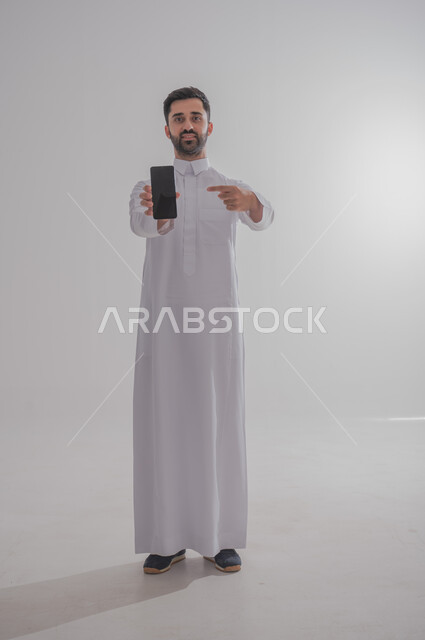 التواصل مع الأهل والأصدقاء عبر الهاتف المحمول، استخدام الأجهزة الحديثة والمتطورة،  بورتريه لرجل عربي خليجي سعودي يرتدي الثوب التقليدي يقوم بعرض شاشة سوداء ، الجسم بالطول الكامل، خلفيه بيضاء