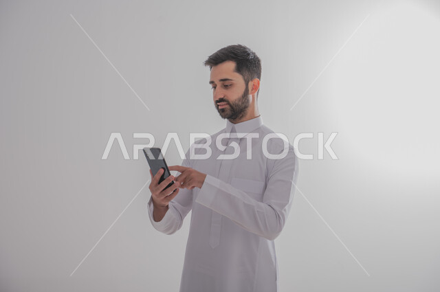 التواصل مع الأهل والأصدقاء عبر الهاتف المحمول، استخدام الأجهزة الحديثة والمتطورة, بورتريه لرجل عربي خليجي سعودي يرتدي الثوب التقليدي يحمل بيده جوال، خلفيه بيضاء