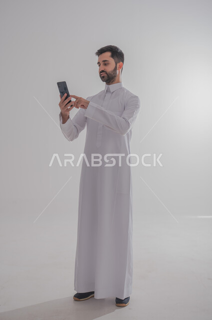 استخدام الأجهزة الحديثة والمتطورة، وسلية للتواصل وتتبع الأخبار، مفهوم الأناقة والرتابة والاهتمام بالمظهر الخارجي, بورتريه لرجل عربي خليجي سعودي يرتدي الزي التقليدي يقوم بالتصفح على الهوتف المحمول، خلفيه بيضاء