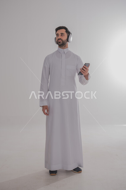 Using advanced technology for electronic digital marketing, displaying modern applications and services, a portrait of a smiling Saudi Gulf Arab young man wearing a traditional dress, wearing headphones on his ears and holding a mobile phone in his hand, full-length body portrait, gray background