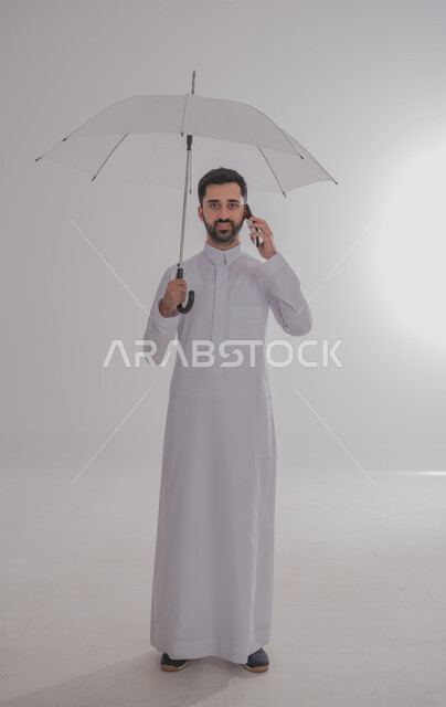 Using advanced technology for digital marketing, displaying modern applications and services, a portrait of a smiling Saudi Gulf Arab young man wearing a traditional dress, holding a mobile phone in one hand and holding an umbrella in the other hand, full-length body portrait, gray background
