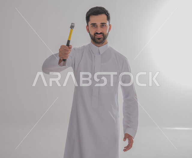 Construction and cladding technician, close-up of smiling Gulf Arab man wearing traditional thobe holding hammer working in engineering sector, looking at camera with gestures of confidence and pleasure, Saudi youth professions and jobs, maintenance work and services, full body image, gray background