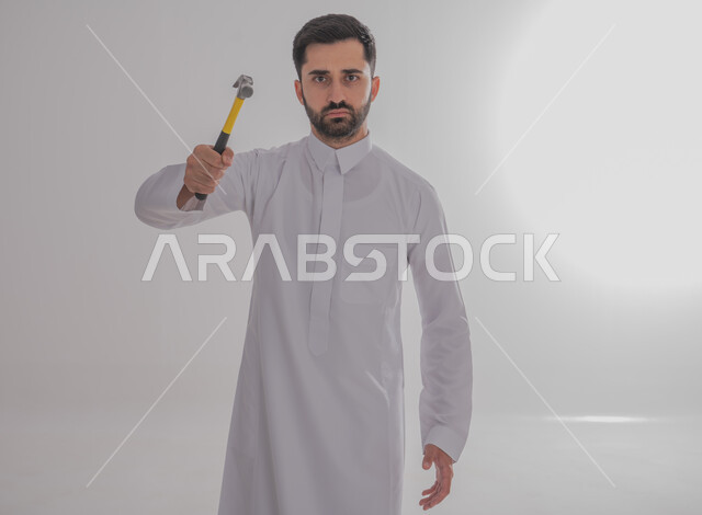 فني بناء وكسوة، لقطة مقربة لرجل خليجي عربي يرتدي ثوبًا تقليديًا ويحمل مطرقة ويعمل في قطاع الهندسة، وينظر إلى الكاميرا بإيماءات غاضبة، مهن ووظائف الشباب السعودي، خدمات الأعمال والصيانة، صورة كاملة للجسم، خلفية رمادية