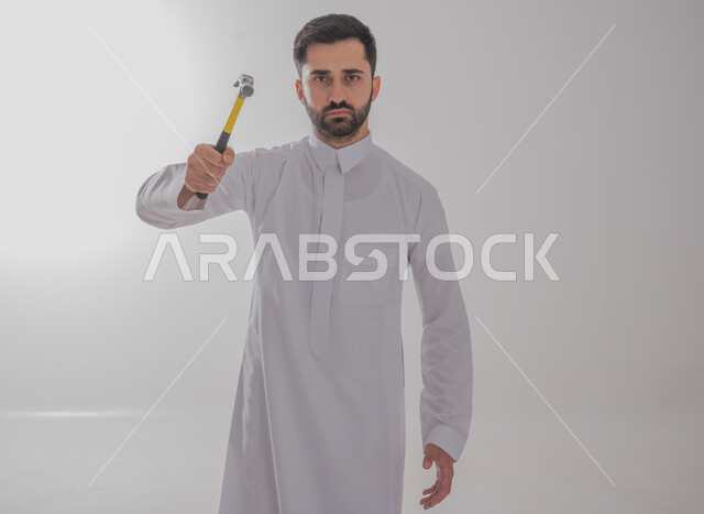 فني بناء وكسوة، لقطة مقربة لرجل خليجي عربي يرتدي ثوبًا تقليديًا ويحمل مطرقة ويعمل في قطاع الهندسة، وينظر إلى الكاميرا بإيماءات غاضبة، مهن ووظائف الشباب السعودي، خدمات الأعمال والصيانة، صورة كاملة للجسم، خلفية رمادية