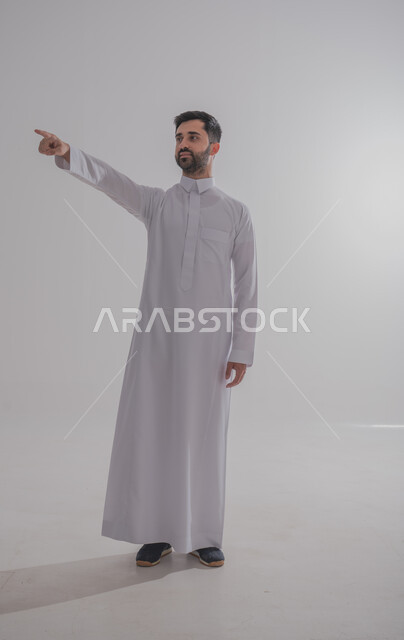 Gestures of affirmation and identification of the corresponding thing, indicating something with the gestures of being here, portrait of a Saudi Gulf Arab man wearing a traditional dress, raising his hands forward with an expression of self-confidence, full-length body portrait, gray background