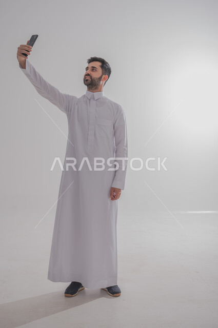 الدردشة مع الأهل والأصدقاء عبر الانترنت، استخدام الأجهزة الحديثة والمتطورة، إجراء مكالمة فيديو عبر وسائل التواصل الاجتماعي، بورتريه لرجل عربي خليجي سعودي يرتدي الثوب التقليدي يقوم بالتقاط صورة سيلفي، الجسم بالطول الكامل، خلفيه بيضاء