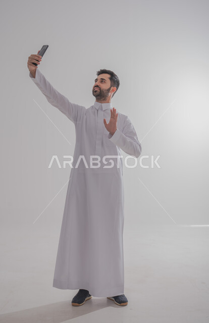 Communicating with family and friends via the Internet, using modern and advanced devices, the concept of elegance, monotony and interest in external appearance, portrait of a Saudi Arabian Gulf man wearing traditional clothing taking a selfie, white background