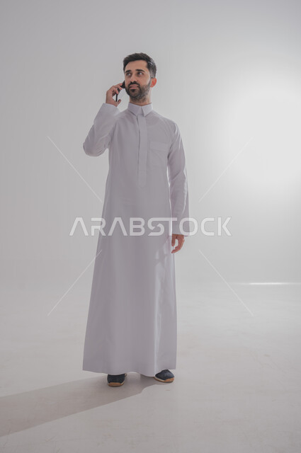 التواصل مع الأهل والأصدقاء عبر الهاتف المحمول، الرد على الاتصالات والمكالمات، استخدام الأجهزة الحديثة والمتطورة،  بورتريه لرجل عربي خليجي سعودي يرتدي الثوب التقليدي يقوم بإجراء مكالمة هاتفية عبر الجوال، الجسم بالطول الكامل، خلفيه بيضاء