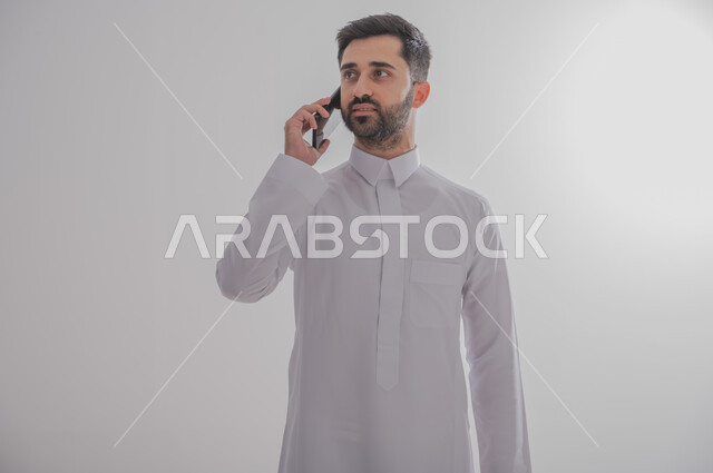 Communicating with family and friends via mobile phone, using modern and advanced devices, answering communications and calls, portrait of a Saudi Gulf Arab man wearing a traditional thobe making a phone call via mobile phone, White background