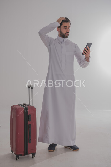 التأخر عن موعد الانطلاق، تعابير الصدمة بتلقي أخبار مزعجة، مفهوم السياحة في إجازة الصيف، الاستعداد لقضاء رحلة صيفية ممتعة، بورتريه لرجل عربي خليجي سعودي يرتدي الزي التقليدي يحمل بيده الهاتف المحمول وبجانبه حقيبة الامتعة، الجسم بالطول الكامل، خلفية بيضاء