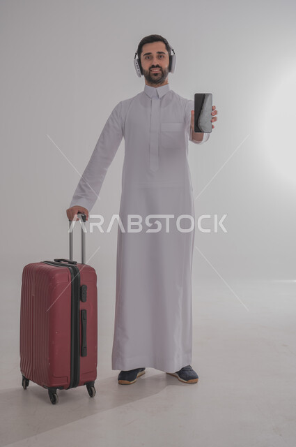 الاستعداد لقضاء رحلة صيفية ممتعة، مفهوم السياحة في إجازة الصيف، القيام بالجولات الترفيهية، بورتريه لرجل عربي خليجي سعودي يرتدي الزي التقليدي وسماعة الرأس يحمل بيده الهاتف المحمول وبجانبه حقيبة الامتعة، عرض شاشة سوداء فارغة، الجسم بالطول الكامل، خلفية بيضاء