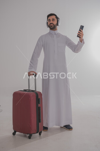 الاستعداد لقضاء رحلة صيفية ممتعة، مفهوم السياحة في إجازة الصيف، القيام بالجولات الترفيهية، بورتريه لرجل عربي خليجي سعودي يرتدي الزي التقليدي وسماعة الرأس يحمل بيده هاتف محمول وحقيبة الامتعة ينظر إلى مكان ما، خلفية بيضاء