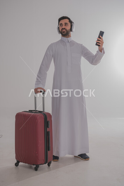 الاستعداد لقضاء رحلة صيفية ممتعة، مفهوم السياحة في إجازة الصيف، القيام بالجولات الترفيهية، بورتريه لرجل عربي خليجي سعودي يرتدي الزي التقليدي وسماعة الرأس يحمل بيده هاتف محمول وحقيبة الامتعة ينظر إلى مكان ما، خلفية بيضاء