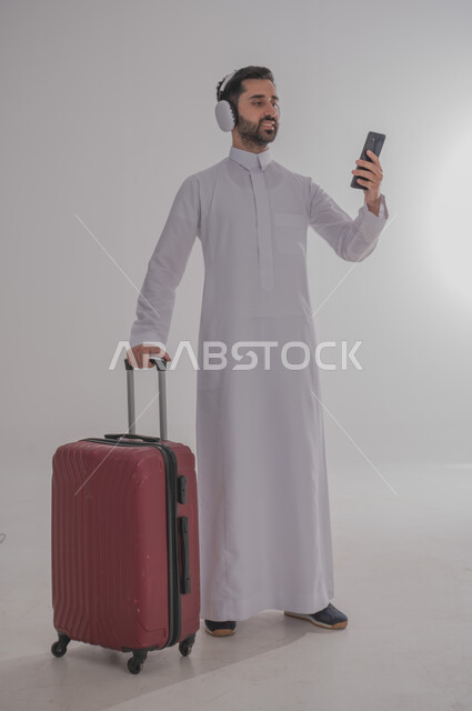 الإتفاق على موعد الانطلاق مع الأصدقاء عبر الانترنت، الاستعداد لقضاء رحلة صيفية ممتعة، مفهوم السياحة في إجازة الصيف، القيام بالجولات الترفيهية، بورتريه لرجل عربي خليجي سعودي يرتدي الزي التقليدي وسماعة الرأس يحمل بيده هاتف محمول وحقيبة الامتعة، خلفية بيضاء