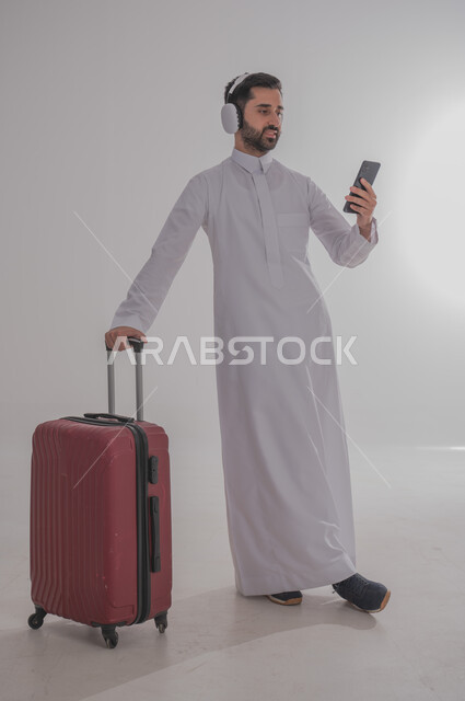 الإتفاق على موعد الانطلاق مع الأصدقاء عبر الانترنت، الاستعداد لقضاء رحلة صيفية ممتعة، القيام بالجولات الترفيهية، بورتريه لرجل عربي خليجي سعودي يرتدي الزي التقليدي وسماعة الرأس يحمل بيده الهاتف المحمول وبجانبه حقيبة الامتعة، الجسم بالطول الكامل، خلفية بيضاء