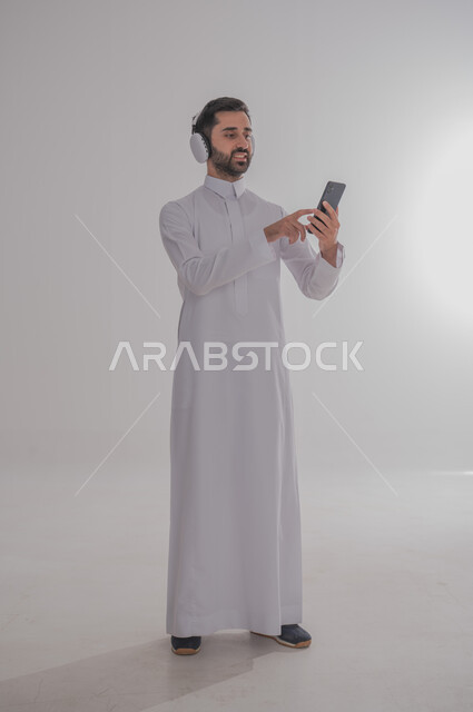 الدردشة مع الأهل والأصدقاء عبر الانترنت، مفهوم الإصغاء والاستمتاع باستخدام الهيدفون، قضاء أوقات ممتعة بالإستماع للمواهب الغنائية، بورتريه لرجل عربي خليجي سعودي يرتدي الثوب التقليدي يضع سماعة الرأس ويحمل الهوتف المحمول، الجسم بالطول الكامل، خلفية بيضاء