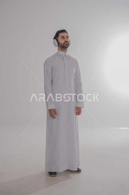 النظر إلى مكان ما بايماءات الطرب والتركيز، مفهوم الإصغاء والاستمتاع بإستخدام الهيدفون، قضاء أوقات ممتعة بالإستماع للمواهب الغنائية، بورتريه لرجل عربي خليجي سعودي يرتدي الثوب التقليدي يضع سماعة الرأس، خلفية بيضاء