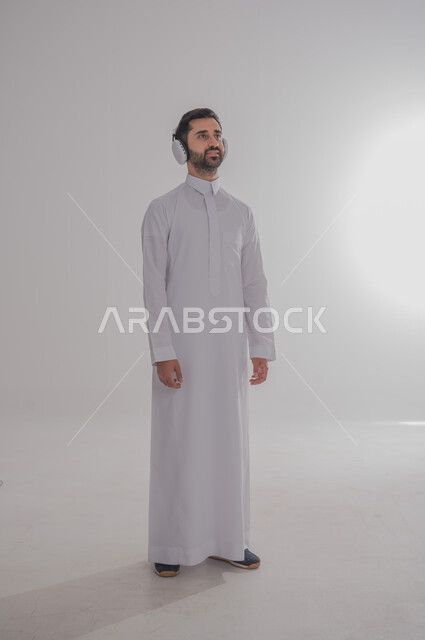 النظر إلى مكان ما بايماءات الطرب والتركيز، مفهوم الإصغاء والاستمتاع بإستخدام الهيدفون، قضاء أوقات ممتعة بالإستماع للمواهب الغنائية، بورتريه لرجل عربي خليجي سعودي يرتدي الثوب التقليدي يضع سماعة الرأس، خلفية بيضاء