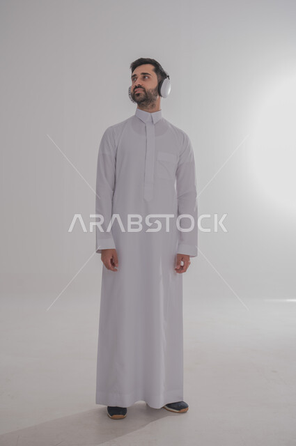 النظر إلى مكان ما بايماءات الطرب والتركيز، مفهوم الإصغاء والاستمتاع بإستخدام الهيدفون، قضاء أوقات ممتعة بالإستماع للمواهب الغنائية، بورتريه لرجل عربي خليجي سعودي يرتدي الثوب التقليدي يضع سماعة الرأس، الجسم بالطول الكامل، خلفية بيضاء