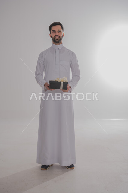 مفهوم الشكر والامتنان، تقديم الهدايا بالاعياد والمناسبات الخاصة، التعبير عن الحب والتقدير والاحترام، بورتريه لرجل عربي سعودي يرتدي الثوب التقليدي يحمل بيديه صندوق هدية أسود اللون وينظر إلى الكاميرا بايماءات السعاده والسرور، الجسم بالطول الكامل، خلفية بيضاء
