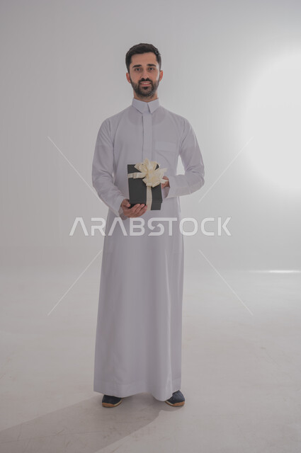 Concept of thanks and gratitude, giving gifts on holidays and special occasions, expressing love, appreciation and respect, portrait of a Saudi Arabian man wearing traditional thobe holding a black gift box in his hands and looking at the camera with gestures of happiness and pleasure, full body, white background