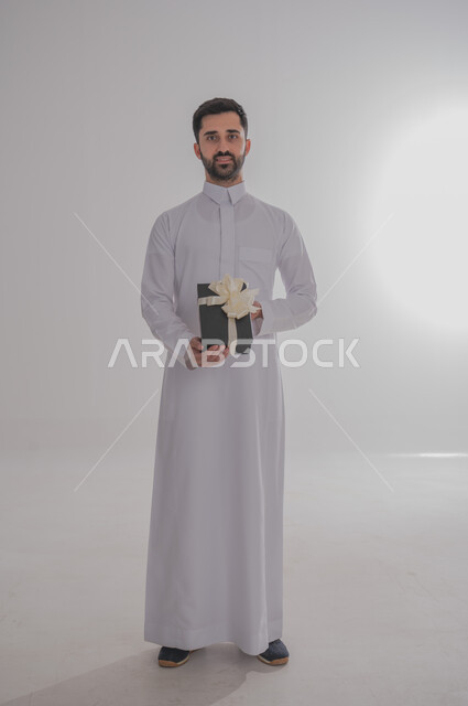 مفهوم الشكر والامتنان، تقديم الهدايا بالاعياد والمناسبات الخاصة، التعبير عن الحب والتقدير والاحترام، بورتريه لرجل عربي سعودي يرتدي الثوب التقليدي يحمل بيديه صندوق هدية أسود اللون وينظر إلى الكاميرا بايماءات السعاده والسرور، الجسم بالطول الكامل، خلفية بيضاء