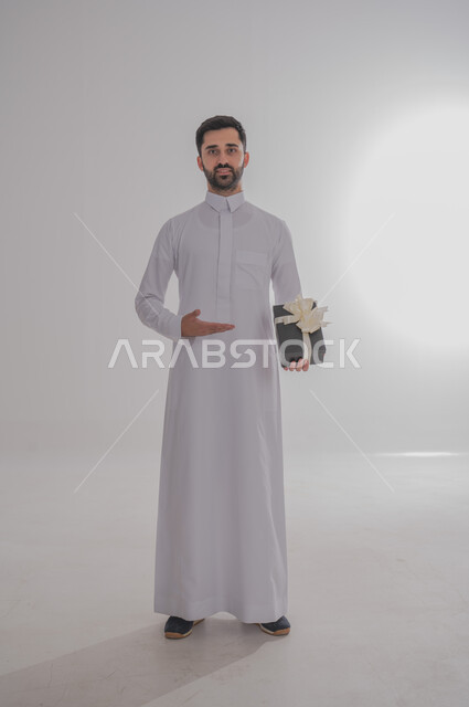 مفهوم الشكر والامتنان، تقديم الهدايا بالاعياد والمناسبات الخاصة، التعبير عن الحب والتقدير والاحترام، بورتريه لرجل عربي خليجي سعودي يرتدي ثوب أبيض يحمل صندوق هدية أسود اللون وينظر إلى الكاميرا بايماءات الابتسامة، خلفية بيضاء 