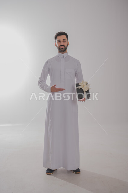مفهوم الشكر والامتنان، تقديم الهدايا بالاعياد والمناسبات الخاصة، التعبير عن الحب والتقدير والاحترام، بورتريه لرجل عربي خليجي سعودي يرتدي ثوب أبيض يحمل صندوق هدية أسود اللون وينظر إلى الكاميرا بايماءات الابتسامة، خلفية بيضاء 