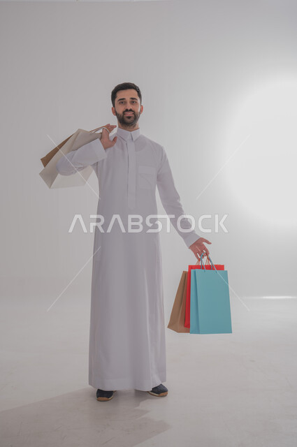 الاستفادة من عروض وتخفيضات الجمعة البيضاء، المتعة في التسوق واقتناء المستلزمات والهدايا، بورتريه لرجل عربي خليجي سعودي مبتسم يرتدي الثوب التقليدي يحمل بيده أكياس ورقية ملونة ينظر إلى الكاميرا بإيماءات السرور والسعادة، الجسم بالطول الكامل، خلفية بيضاء