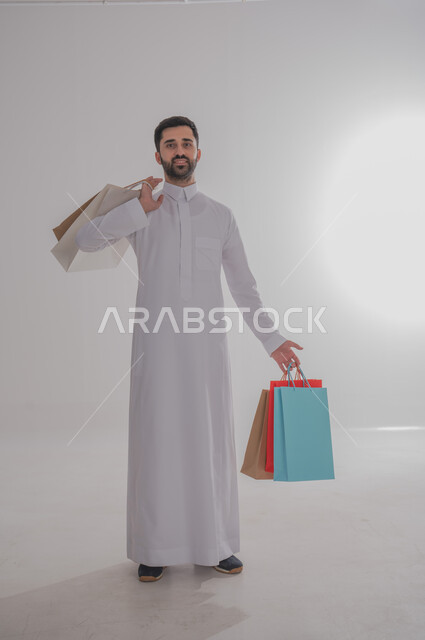 الاستفادة من عروض وتخفيضات الجمعة البيضاء، المتعة في التسوق واقتناء المستلزمات والهدايا، بورتريه لرجل عربي خليجي سعودي مبتسم يرتدي الثوب التقليدي يحمل بيده أكياس ورقية ملونة ينظر إلى الكاميرا بإيماءات السرور والسعادة، الجسم بالطول الكامل، خلفية بيضاء