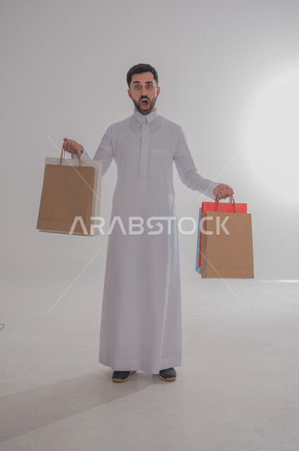 التفاجؤ من شيء ما، الاستفادة من عروض وتخفيضات الجمعة البيضاء، اقتناء المستلزمات والهدايا، بورتريه لرجل عربي خليجي سعودي يرتدي الثوب التقليدي يحمل بيده أكياس تسوق ورقية ملونة ينظر إلى الكاميرا بإيماءات الدهشة والتعجب، الجسم بالطول الكامل، خلفية بيضاء