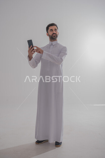 Communicating with family and friends via mobile phone, using modern and advanced devices, portrait of a Saudi Gulf Arab man wearing a traditional thobe, holding a mobile phone in his hand, white background