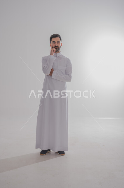 النظر إلى الكاميرا بايماءات التفكير والشرود، مفهوم الأناقة والاهتمام بالمظهر الخارجي، بورتريه لرجل عربي خليجي سعودي يرتدي الثوب الابيض يضع يده على ذقنه بايماءات التخطيط والتركيز، صورة الجسم بالطول الكامل، خلفية بيضاء 