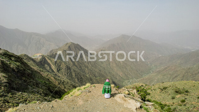 تصوير درون من اعلى لرجل عربي خليجي سعودي يرتدي الزي الخليجي التقليدي ، يضع على ظهره علم المملكة العربية السعودية ، ذكرى اليوم الوطني السعودي ، الاحتفال بالعيد الوطني السعودي على جبل السودة في ابها جنوب المملكة العربية السعودية