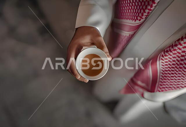 منتج وطني سعودي محلي، إكرام الضيف و حسن الضيافه والاستقبال, تقديم المشروبات الساخنة اللذيذة, صورة مقربة ليدين رجل عربي سعودي خليجي يرتدي الثوب والشماغ السعودي التقليدي يمسك بيده فنجان القهوة