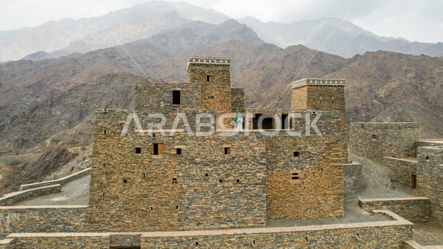 Drone photography from afar of a Saudi Arabian Gulf man wearing traditional Saudi dress, standing on one of the ancient houses in the ancient village of Dhi Ayn in Al-Baha, southern Saudi Arabia, holding the flag of the Kingdom of Saudi Arabia, the annive