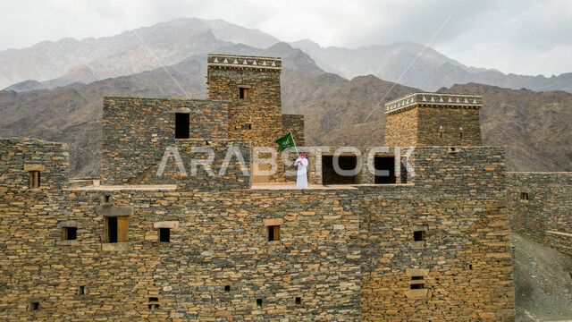 Drone photography from afar of a Saudi Arabian Gulf man wearing traditional Saudi dress, standing on one of the ancient houses in the ancient village of Dhi Ayn in Al-Baha, southern Saudi Arabia, holding the flag of the Kingdom of Saudi Arabia, the annive