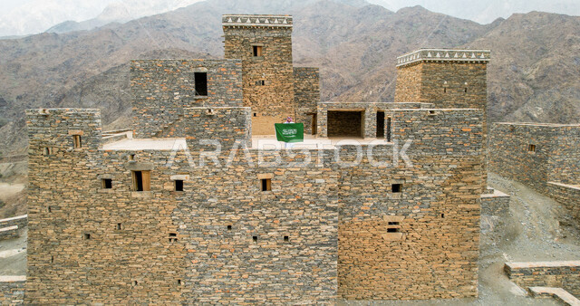 تصوير درون من بعيد لرجل عربي خليجي سعودي يرتدي الزي التقليدي السعودي ،  يقف على احد البيوت الاثرية في قرية ذي عين الاثرية في الباحة جنوب السعودية ، بيده علم المملكة العربية السعودية ،ذكرى اليوم الوطني السعودي ، الاحتفال بالعيد الوطني السعودي