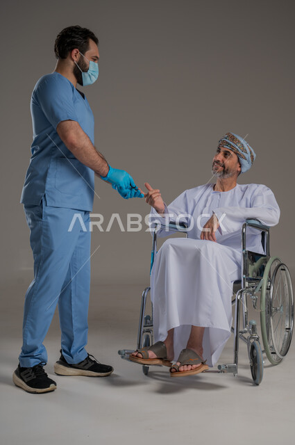 ايماءات السرور والسعادة، مفهوم العناية ورعاية المرضى والمعاقين، بورتريه لممرض يرتدي اليونيفورم والكمامة يقوم بالاعتناء برجل عربي خليجي عماني من ذوي الاحتياجات الخاصة يرتدي الدشداشة والعمامة يجلس على كرسي متحرك، خلفية رمادية