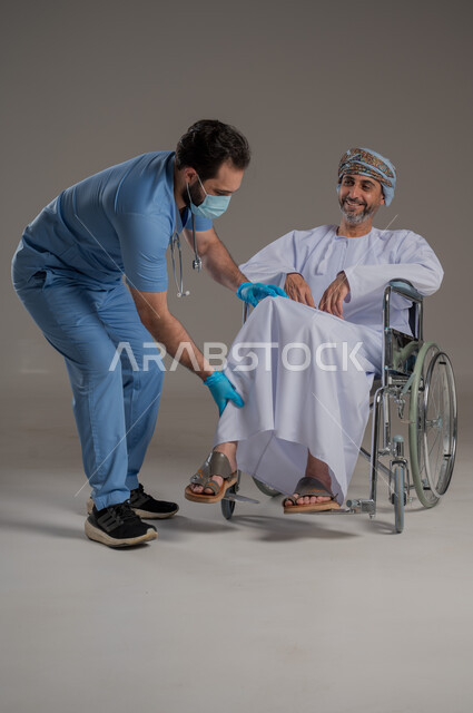 ايماءات السرور والسعادة، مفهوم العناية ورعاية المرضى والمعاقين، بورتريه لممرض يرتدي اليونيفورم والكمامة يقوم بالاعتناء برجل عربي خليجي عماني من ذوي الاحتياجات الخاصة يرتدي الدشداشة والعمامة يجلس على كرسي متحرك، خلفية رمادية