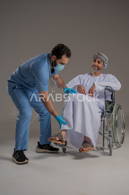 ايماءات السرور والسعادة، مفهوم العناية ورعاية المرضى والمعاقين، بورتريه لممرض يرتدي اليونيفورم والكمامة يقوم بالاعتناء برجل عربي خليجي عماني من ذوي الاحتياجات الخاصة يرتدي الدشداشة والعمامة يجلس على كرسي متحرك، خلفية رمادية