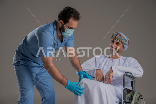 ايماءات السرور والسعادة، مفهوم العناية ورعاية المرضى والمعاقين، بورتريه لممرض يرتدي اليونيفورم والكمامة يقوم بالاعتناء برجل عربي خليجي عماني من ذوي الاحتياجات الخاصة يرتدي الدشداشة والعمامة يجلس على كرسي متحرك، خلفية رمادية