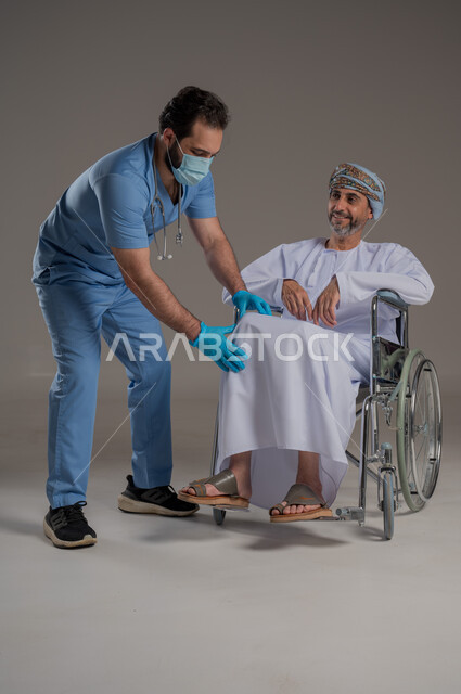 ايماءات السرور والسعادة، مفهوم العناية ورعاية المرضى والمعاقين، بورتريه لممرض يرتدي اليونيفورم والكمامة يقوم بالاعتناء برجل عربي خليجي عماني من ذوي الاحتياجات الخاصة يرتدي الدشداشة والعمامة يجلس على كرسي متحرك، خلفية رمادية